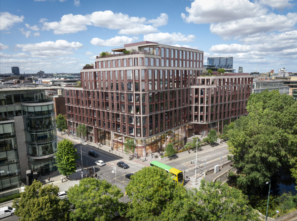 Modern multi-storey glass and brick office building at a busy street corner, surrounded by trees and traffic.
